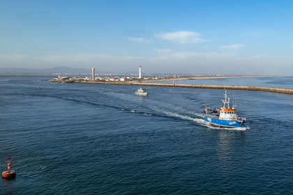 Portugal, Algarve, Ria Formosa Natural Park, Faro, fishing boat leaving the port, the lighthouse of Ilha do Farol part of Ilha da Culatra in the background (aerial view)