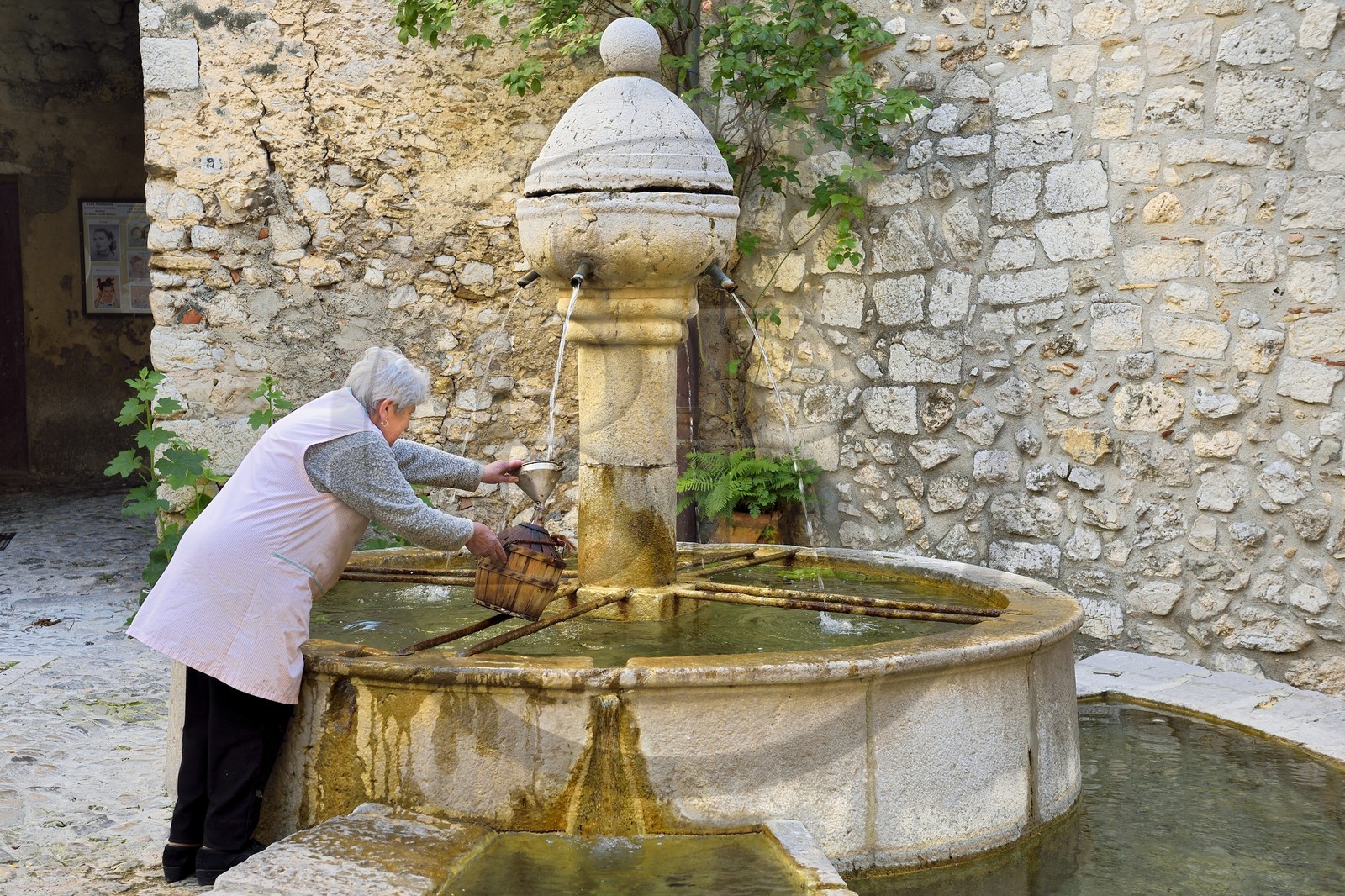 France, Alpes-Maritimes (06), Peille, femme remplissant une bonbonne à la fontaine place du Mont Agel